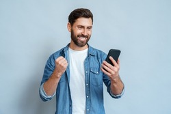 Happy satisfied man looking at mobile phone screen gesturing yes with clenched fist isolated on blue studio copy space. Overjoyed excited joyful guy make winner gesture read message about reward