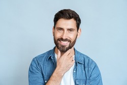 Headshot portrait of thinking stylish young adult caucasian man smiling and touching beard on face looking at camera standing over blue studio wall background. Different people emotion and expression