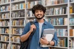 Portrait of cheerful male international Indian student with backpack, learning accessories standing near bookshelves at university library or book store during break between lessons. Education concept