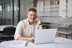 Middle aged Latin or Indian businessman having call on smartphone with business partners or clients. Smiling mature Hispanic man sitting at table talking by mobile cellphone at workplace in office. 
