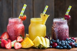 Summer Slushies from Blueberries, Cherries, Lemon, Mango, Strawberries, Lime, Pineapple and Ice with Ingredients nearby, on dark rustic background