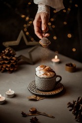 Hot cappuccino coffee mug on a table decorated with milk foam, candles. Holiday Merry Christmas mood food photo. Cinnamon falling in the cup