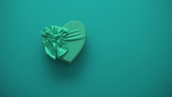 Green gift box on a dark cyan green background. Heart-shaped gift box with a ribbon. Happy Valentine's Day. Anniversary. Top view flat lay space for text. Minimal luxury.