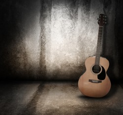 An wooden acoustic guitar is against a grunge textured wall. The room is dark with a spotlight for your copyspace. Use it for a music or concert concept.