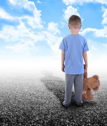 A young boy is holding a teddy bear and standing on an empty road with clouds in the sky. His shadow is in the horizon.