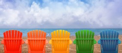 A rainbow of colors of wooden beach chairs are lined up along the water shore. There is copyspace in the clouds for a vacation message.