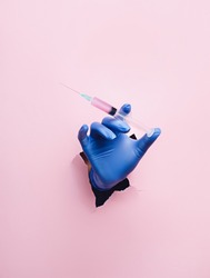 Doctor hand with surgical gloves breaking through the wall and holding syringe on pink background. Creative mass vaccination concept. Covid-19, influenza or flu global pandemic immunization.