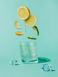Slices of lemons and lime flying into the glass with ice cubes and water isolated on a pastel mint blue background. Ingredients for a mojito cocktail. Creative refreshing summer drink concept.