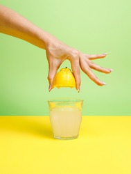 A woman's hand squeezing half of a lemon and making a glass of icy cold lemonade isolated on a yellow and mint green background. Cocktail ingredient. Creative summer refreshing drink concept