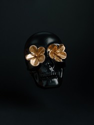 Black glossy 3D human skull with two golden flowers instead of eyes isolated on black background. Creative Halloween or Santa Muerte concept. Minimal dark cult aesthetic.