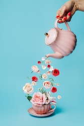 Creative layout with hand holding teapot and pouring fresh flowers and leaves into tea cup on pastel blue background. Creative floral spring bloom concept. Still life natural visual trend. 