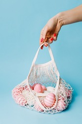 Woman's hand holding crochet bag full of colorful eggs against pastel blue background. Minimal Easter holiday concept. Spring holiday shopping, advertisement or sale banner idea.