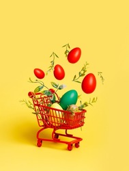 Shopping trolley with flying colored eggs on vibrant yellow background. Easter celebration or delivery concept with copy space. Sale and traditional spring shopping for the holidays.