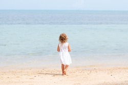 A little girl with curly hair in a white dress walks along a sandy beach on the shore of the sea, ocean and smiles. Sea holidays, travel and beach holidays with children
