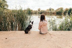 Little girl sitting with a Boston Terrier dog on the lakeshore - with her back turned, her dog staring at water - Autumn scenery