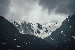 Dark atmospheric mountain landscape with glacier on black rocks in lead gray cloudy sky. Snowy mountains in gray low clouds in rainy weather. Gloomy mountain landscape with rocky mountains with snow.