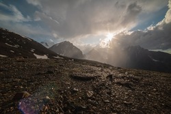 Dramatic alpine landscape with sunlit mountain with sharp stones with view to high snow mountains and large glacier. Bright sun in overcast sky above snowy mountain range. Stones shining in sunlight.
