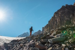 Scenic alpine landscape with silhouette of hiker with trekking poles against large sharp rocks and snow mountain peak in sunlight. Man with backpack in high mountains under blue sky in sunny day.
