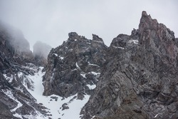 Gloomy mountain landscape with high sharp rockies with snow in gray low clouds. Closeup of sharp rocks and peaked tops in gray cloudy sky. Bleak overcast scenery with mountain range with pointy rocks.
