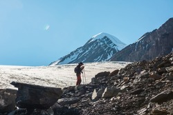 Scenic alpine landscape with silhouette of hiker with trekking poles against large glacier tongue and snow mountain peak in sunlight. Man with backpack in high mountains under blue sky in sunny day.
