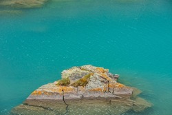 Stone with grass and lichens in azure transparent water of mountain lake. Fragment of meditative lake with lichen rock. Scenic nature background of relaxing shiny ripples on calm water of alpine lake.