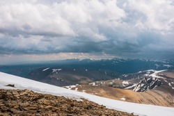 Dramatic alpine view from stone hill with snow to snowy mountain top in light under cloudy sky. Atmospheric landscape at high altitude with cloudiness. High snow mountain peak in sunlight in overcast.