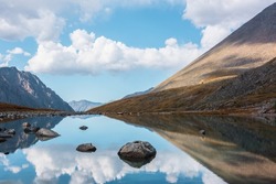 Tranquil autumn landscape with clouds reflection on smooth mirror surface of mountain lake in high hanging valley. Meditative view from calm alpine lake to mountain vastness. Stones in clear water.