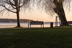 Empty park bench in front of an idyllic lake. The sun is going down and the sky is glowing in orange color. Tranquil scenery without people. Landscape with a resting place in a lakeside area.