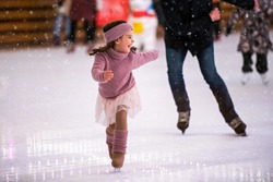 Laughing little girl in a pink sweater is skating on a winter evening on an outdoor ice rink, it is snowing
