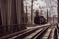 an old vintage steam locomotive with huge clouds of smoke rides along the railway among the wires, poles and iron supports of the bridge