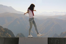 Young woman balancing on the edge of an abyss walks along the road parapet against the backdrop of mountains. Risk, black and white streak of life concept.