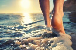 Closeup of a man's bare feet walking at a beach at sunset, with a wave's edge foaming gently beneath them, toned colors