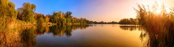 Panorama of a gorgeous sunset at a lake, with gold and blue color and trees reflected in the water
