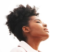 Beautiful young majestic black woman wearing a light pink jacket with a high puff updo hairstyle holds her head back against a white background outside with eyes closed                              