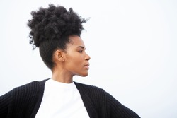 Beautiful, regal, young black woman stands powerfully with her arms outstretched, wearing a white shirt, black sweater, and an updo hairstyle, looking right against sky                               
