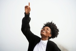 Beautiful young black woman points up to the heavens and god with her eyes closed and smiling outside in the fog at the ocean                               