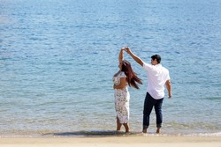 Beautiful young couple in love at the beach dancing and twirling at the blue water's edge on a sunny morning                               