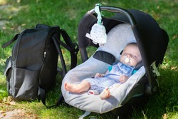 Little baby on vacation road trip sitting in baby car seat on the grass with backpack :  concept of safe travel and transportation of babies during vacation journeys.