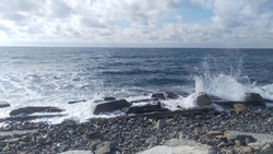 The foamy sea wave hits the rocky shore. The surf hits the concrete blocks lying on the shore and splashing. The sky above the sea is blue with clouds