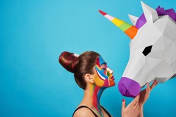 Sideview of pop art portrait of model wearing black opened top. Girl has saturated make up with bright geometrical figures and fancy hairdress. Posing on blue background with pink paper unicorn's head