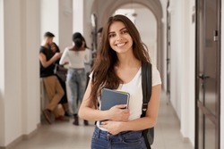 Positivity beautiful girl smiling at camera, standing on corridor with notes as backpack, going to lesson. Happy brunette female student studying in luxury university.