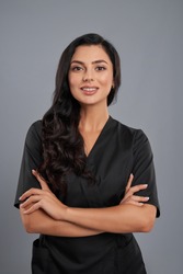 Attractive female beautician in black uniform standing with crossed arms over grey background. Positive cosmetologist with long dark hair smiling and looking at camera.