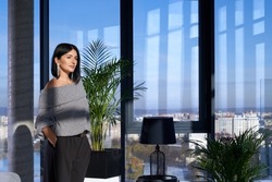 Side view of pensive beautiful woman fashionable dressed standing near large panoramic window and enjoying view on city. Concept of modern house with stylish interior and window.