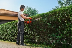 Strong caucasian man in uniform, safety gloves and mask pruning hedge with petrol trimmer. Male gardener shaping green overgrown bushes outdoors.