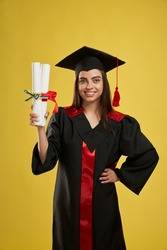 Front view of pretty female graduate standing, holding two diplomas. Student with bachelor, master degree smiling, looking at camera, happy. Isolated on yellow studio background.