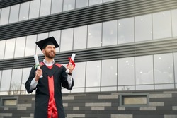 Front view of student with beard graduating from college, getting degree. Male wearing graduate gown and cap, standing, holding diploma, smiling, happy. Concept of education.