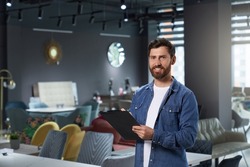 Successful store owner in denim shirt making notes, while working in modern furniture shop. Portrait of handsome designer with tablet smiling at camera, while standing in expo center. Concept of work.