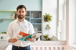Smiling male sales manager holding catalogue of various furniture, while standing in light showroom. Portrait of bearded customer flipping brochure, while choosing goods. Concept of interior design.