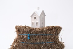 A small ceramic house on a weak foundation of a bale of hay. Great for concepts about foundations, home purchasing, insurance, or other abstract ideas. Horizontal with copy space.