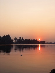 A red and orange Indian sunset reflects over a river in Kerala with palm trees in silhouette in the background